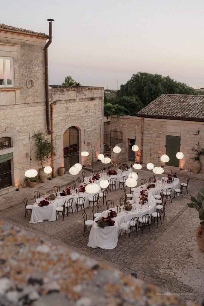 Serpentine tables with drapery, deep red, burgundy florals and lantern lighting for Italian wedding at Villa Carcara in Sicily.